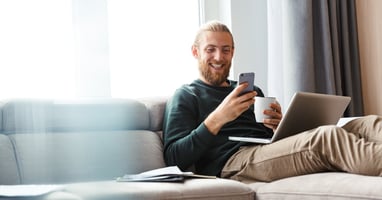 A smiling young man lies on a couch, holding a mug, and looking at his phone. An open laptop sits on his lap.