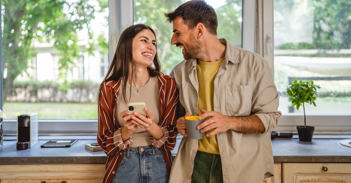 A man and a woman stand in the kitchen smiling at each other. She's holding a cell phone, and he's holding a mug.