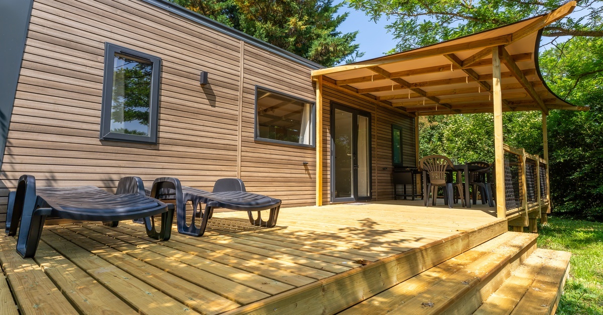 A manufactured home features a large, wooden deck with a tan awning, an outdoor dining set, and two lounge chairs.