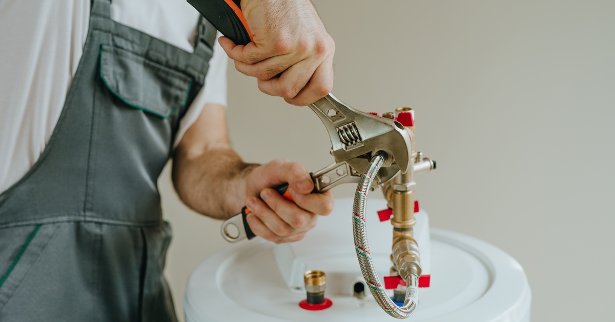 A close-up view shows a person wearing gray overalls using a wrench on a metal tube that attaches to a water heater.