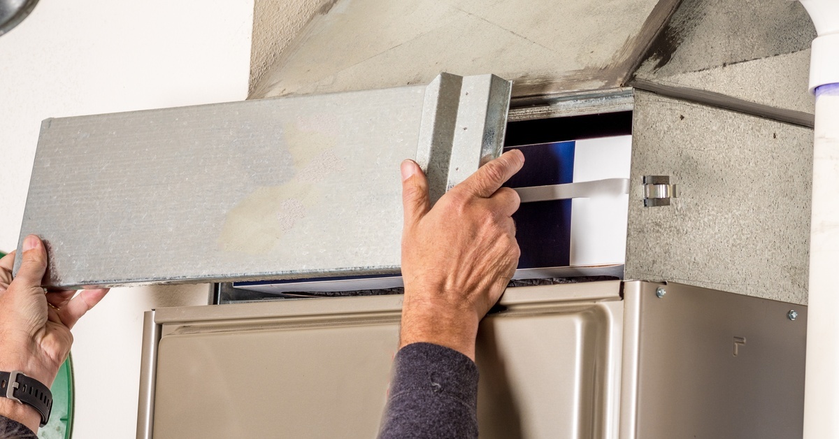 A person wearing a gray long-sleeve shirt and glasses is pulling off a metal door on the furnace.