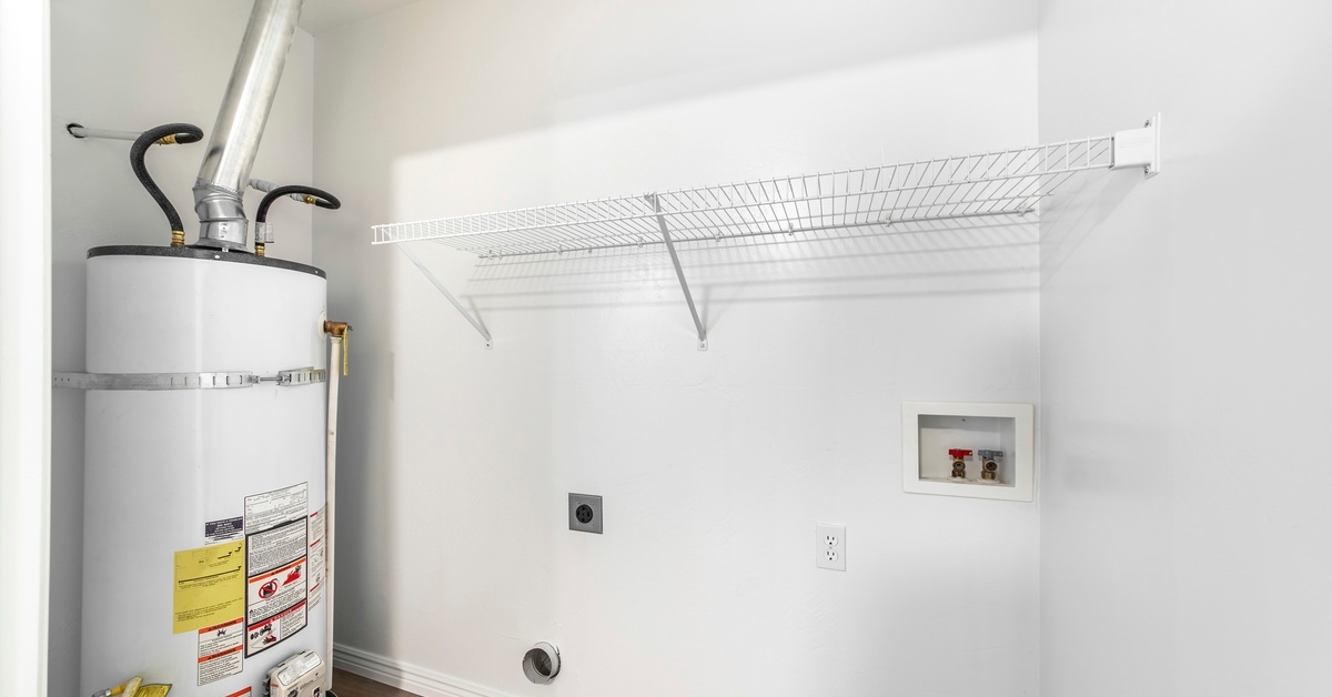 A white hot water heater sits inside a utility room with white walls, laundry hookups, and a white wire shelf.