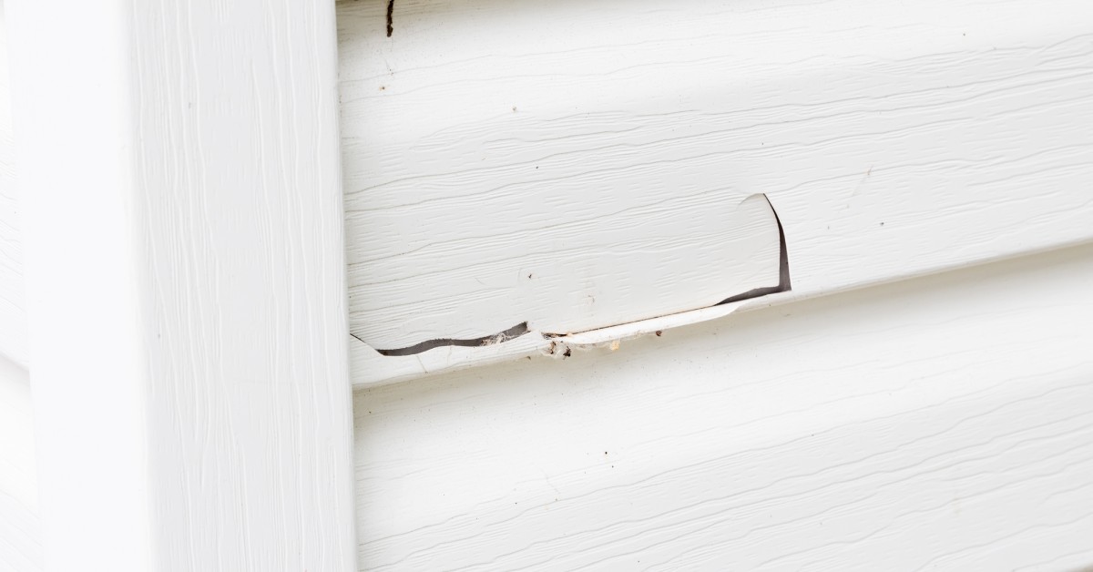 A close-up view shows a large horizontal crack on a home's white siding, exposing the insulation underneath.