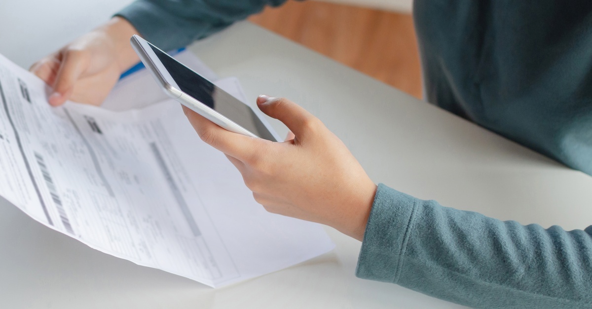 A close-up view shows a person sitting at a white table holding their phone in one hand and a bill in the other.