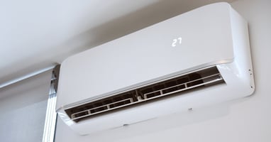A close-up view shows a white, ductless air conditioning unit attached to the white wall of a home.