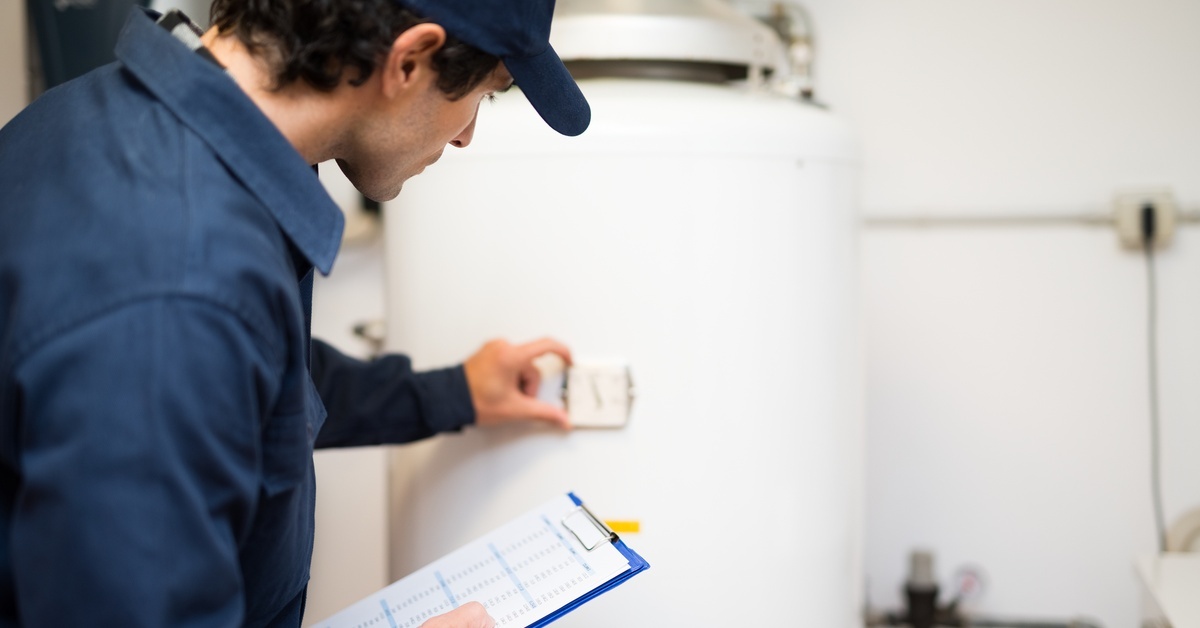 A technician wearing a blue jumpsuit and a blue hat holds a device up to a white water heater in a home.