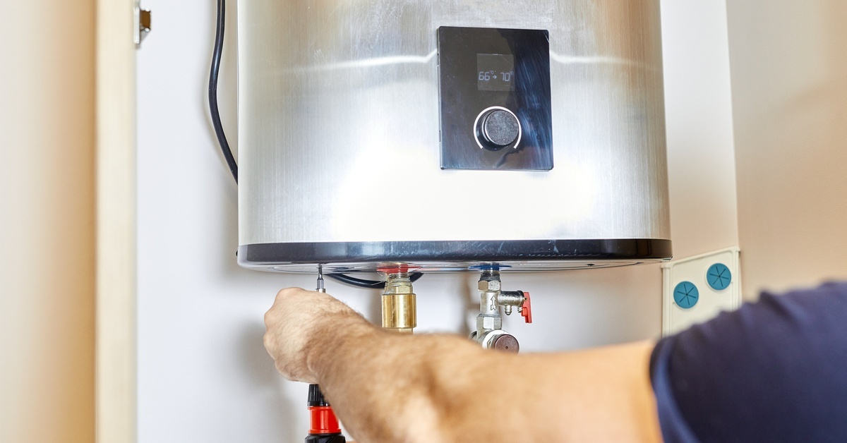 A close-up view shows a person using a black and red screwdriver to tighten the screw on a water heater.