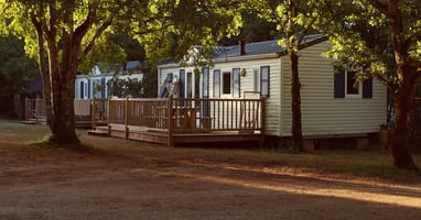 Two mobile homes sit side by side on a piece of wooded property. One has yellow siding and one has white siding.
