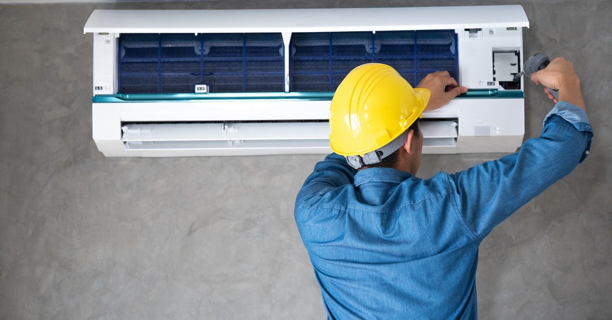 A technician wearing a blue long-sleeve shirt and a yellow hard hat is working on a mini split AC system.