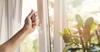 A close-up view shows a person using their hand to open a window with white trim. Sunshine is coming through the window.