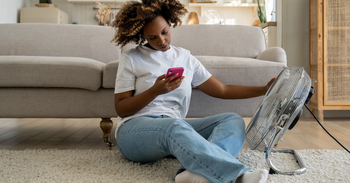 A woman sits on the floor on a rug in front of a gray couch. She's scrolling on her phone with an arm resting on a fan.