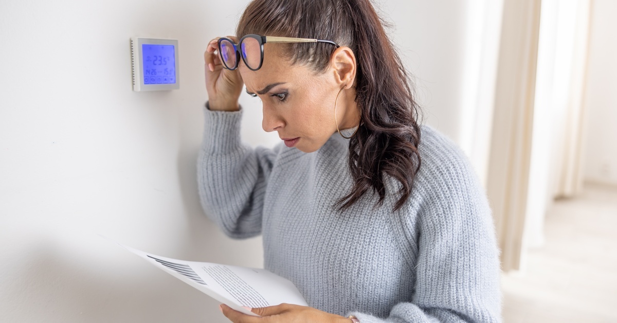 A woman wearing a gray sweater holds a piece of paper and lifts up her glasses to look at it. She looks concerned.