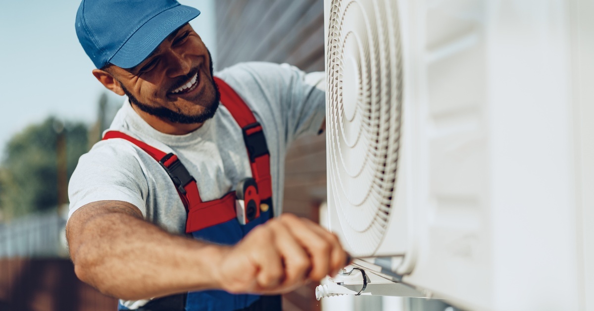 A smiling man wearing a white T-shirt and red overalls is working on a white air conditioning unit outside.