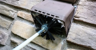 A close-up view of a person using a large, bristled brush to clean an air duct on the home's exterior.