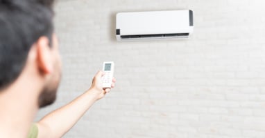 A man with brown hair holding a remote and pointing it at a split system that’s on a white brick wall.