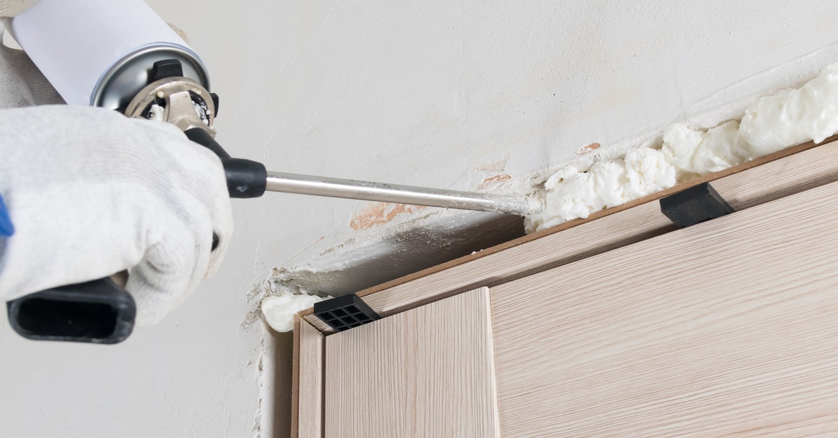 A gloved hand uses a spray foam tool to apply white spray foam inside a gap along an entryway.