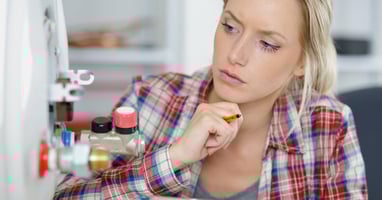 A woman holds a clipboard and touches her chin as she looks closely at her home water heater valves.