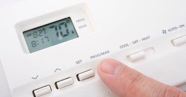 A finger presses the "Program" button on a white thermostat with a small digital screen that reads 70 degrees.