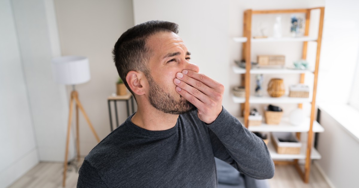 A person wearing a black long-sleeve shirt stands in a living room holding his nose. He has a disgusted look on his face.