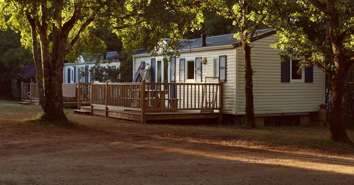 Two mobile homes sit side by side on a piece of wooded property. One has yellow siding and one has white siding.