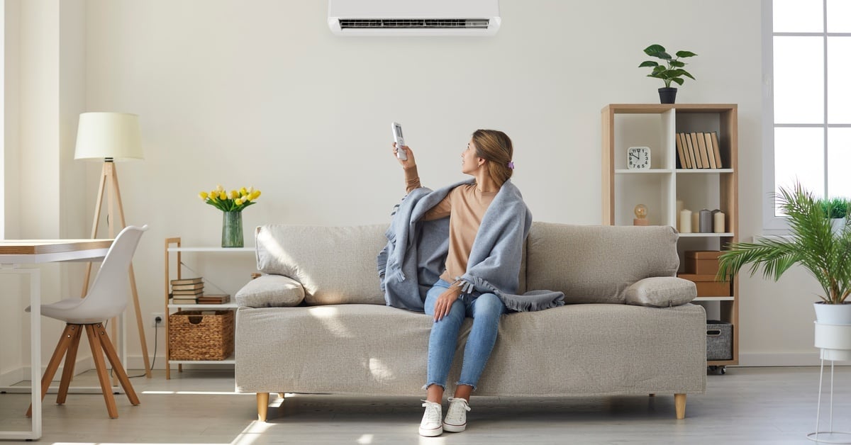 A woman sitting on a couch with a blanket draped over her shoulders is pointing a remote at a mini split AC unit on the wall.