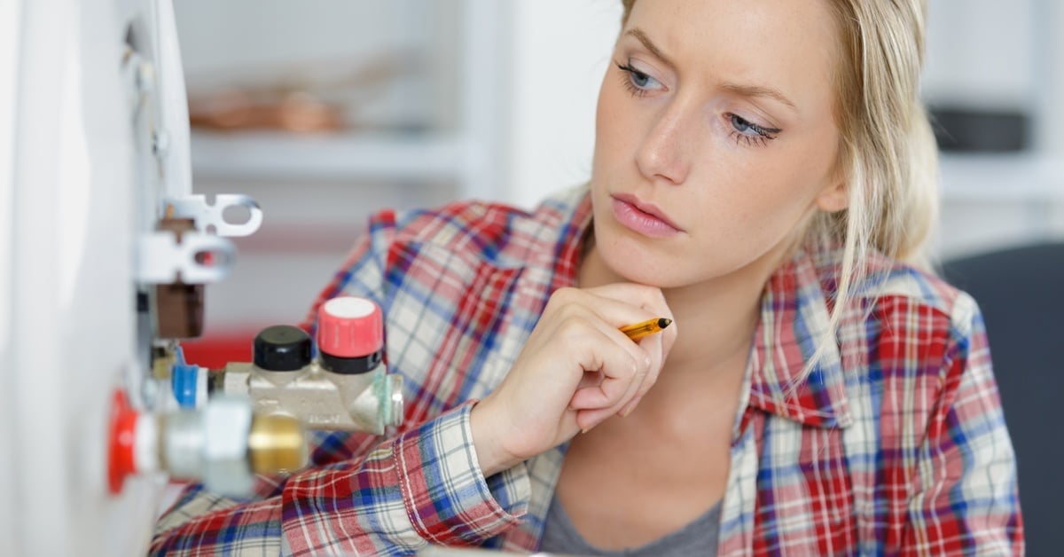 A woman holds a clipboard and touches her chin as she looks closely at her home water heater valves.